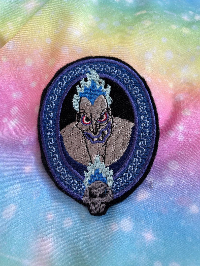 May include: An embroidered iron-on patch featuring a purple and blue cartoon image of the Greek god Hades from Disney's Hercules. The image is surrounded by a blue and purple circular frame.