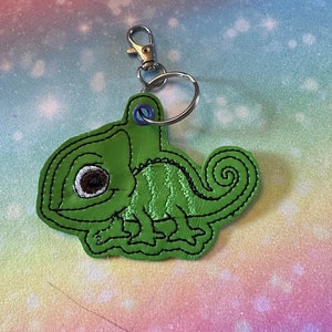 May include: A green embroidered chameleon keyring with a black outline and a brown eye. The keyring is attached to a silver split ring.
