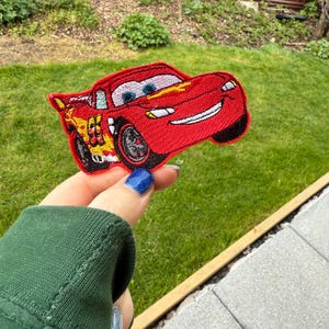 Puede incluir: Un parche bordado rojo con Rayo McQueen de la película Cars. El parche muestra el coche con una cara sonriente, neumáticos negros y el número 95. El parche se encuentra sobre un fondo verde.