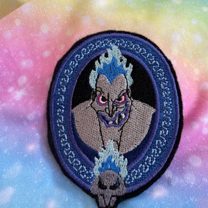 May include: An embroidered iron-on patch featuring a cartoon image of Hades from Disney's Hercules. Hades is depicted with his signature blue flames and a menacing expression. The patch is surrounded by a blue and white decorative border.