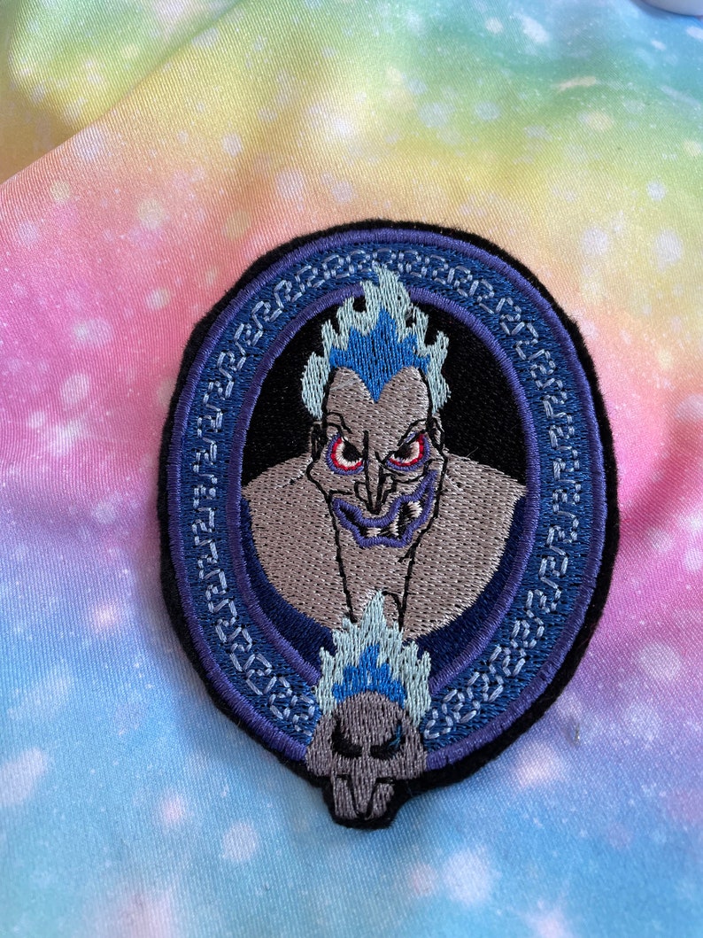 May include: An embroidered iron-on patch featuring a cartoon image of Hades from Disney's Hercules. Hades is depicted with his signature blue flames and a menacing expression. The patch is surrounded by a blue and white decorative border.