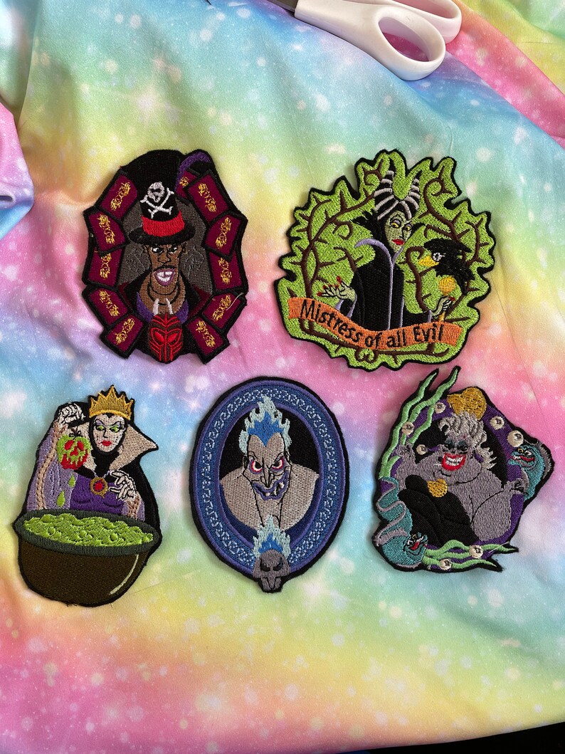 May include: Five embroidered iron-on patches featuring Disney villains. The patches are on a rainbow-colored fabric background. The villains are Ursula, Hades, Maleficent, Evil Queen, and Captain Hook.