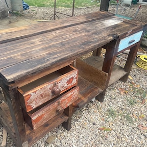 May include: A rectangular wooden workbench featuring a dark brown stained top and frame. It includes two stacked drawers on the left with distressed orange paint and round wooden knobs, and one light blue drawer on the right with a dark handle.