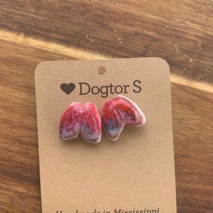 May include: A pair of anatomical lung-shaped earrings in red and white, displayed on a brown card. The card has the text "Dogtor S" with a heart symbol and "Handmade in Mississippi". The earrings are a unique accessory.