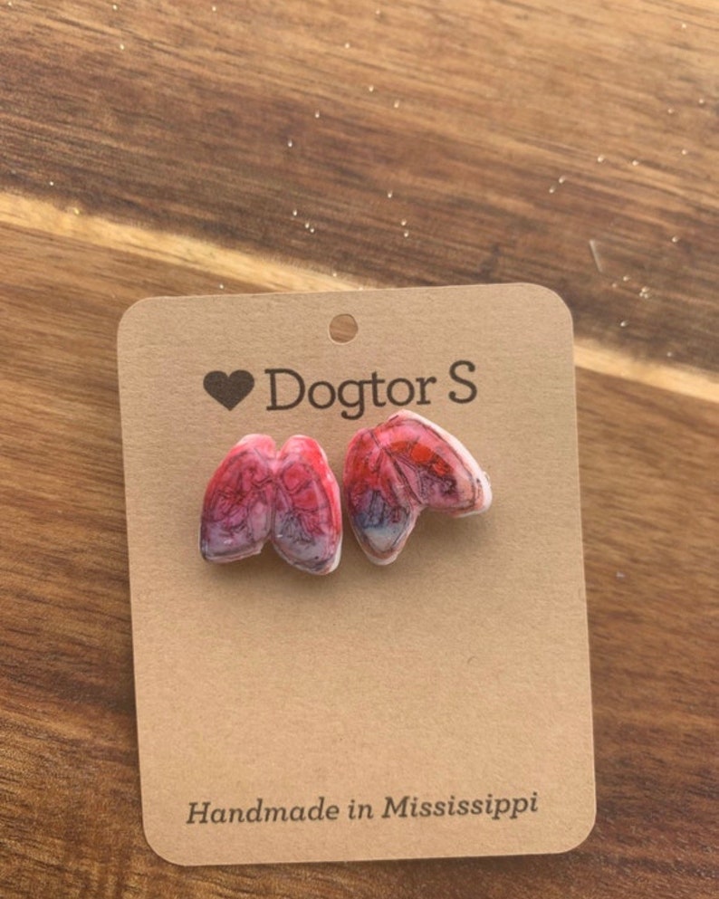 May include: A pair of anatomical lung-shaped earrings in red and white, displayed on a brown card. The card has the text "Dogtor S" with a heart symbol and "Handmade in Mississippi". The earrings are a unique accessory.
