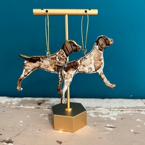 Pode incluir: Um par de brincos de argola dourados com a silhueta de um cão castanho e branco. Os brincos são exibidos em um suporte dourado.