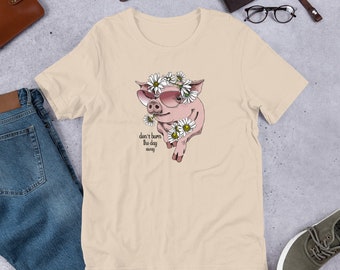 May include: A light beige t-shirt featuring a pink pig wearing sunglasses and a daisy flower crown. The text "don't burn the day away" is printed on the shirt.