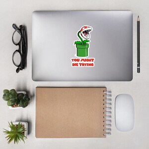 May include: A sticker featuring a cartoon Piranha Plant from the Super Mario video game series. The plant has a red and white head, green stem, and is in a green pipe. The text "YOU MIGHT DIE TRYING" is in red.