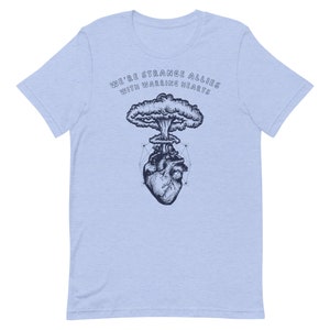 May include: A light blue t-shirt with a graphic of a heart with a mushroom cloud exploding from it. The text "We're strange allies with warring hearts" is printed on the shirt.