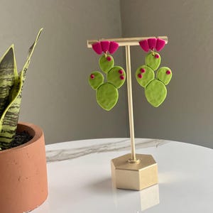 May include: A pair of handmade cactus-shaped earrings. The earrings feature bright green, textured cactus pads with small pink dots and vibrant fuchsia flower accents. They are displayed on a gold-colored earring stand.