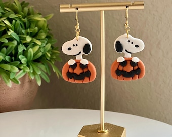 Puede incluir: Un par de pendientes colgantes con un perro de dibujos animados blanco sosteniendo una calabaza naranja con una cara de calabaza negra. Los pendientes están colgados de un soporte dorado.