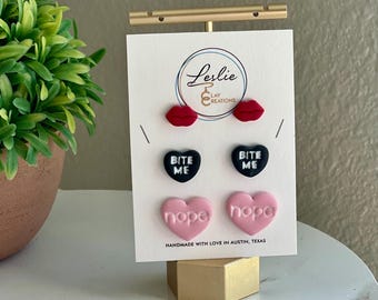 May include: A set of three pairs of polymer clay earrings displayed on a white card with the text "Leslie Lay Creations" and "Handmade with love in Austin, Texas". The earrings are shaped like lips, hearts, and candy hearts. The lips are red, the hearts are black with white text that says "Bite Me", and the candy hearts are pink with white text that says "nope".