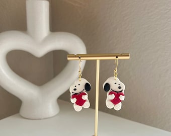 May include: A pair of handmade earrings featuring a white cartoon dog with black ears and a red heart. The earrings are suspended from gold-colored hooks and displayed on a gold-colored earring stand.
