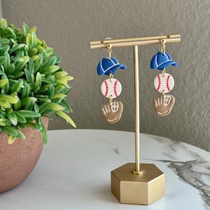 Könnte beinhalten: Ein Paar Hängeohrringe mit einem Baseball, einem Baseballhandschuh und einer Baseballkappe. Die Ohrringe sind aus Polymer Clay gefertigt und haben eine goldene Oberfläche.