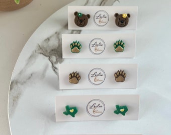 May include: A collection of handmade stud earrings featuring various designs. The earrings include bear faces, paw prints, Texas state shapes, and Baylor University logos. Each pair is displayed on a white card with the maker's logo.