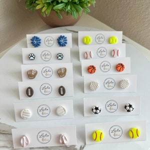 Op de afbeelding: Een set van 10 paar polymer clay oorbellen met verschillende sportthema-ontwerpen, waaronder honkballen, basketbals, voetbals, volleybals, voetbals, golfballen en meer. De oorbellen worden gepresenteerd op witte kaartjes met de merknaam "Leslie & Creations" gedrukt op elk kaartje.