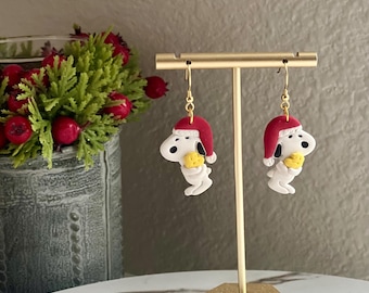 Puede incluir: Un par de pendientes colgantes con un perro de dibujos animados blanco que lleva un gorro de Papá Noel rojo y sostiene un hueso amarillo. Los pendientes están colgados de un soporte dorado.