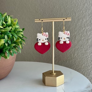 Puede incluir: Un par de pendientes en forma de corazón rojo con un personaje de Hello Kitty blanco sosteniendo el corazón. Los pendientes están en un soporte dorado.