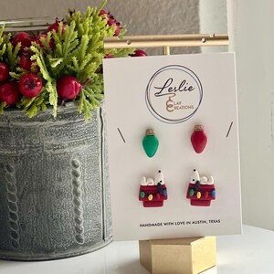 Puede incluir: Un conjunto de tres pares de pendientes de arcilla polimérica con luces de Navidad y un perro en una caseta. Los pendientes se muestran en una tarjeta blanca con el texto "Leslie Clay Creations" y "Handmade with love in Austin, Texas".