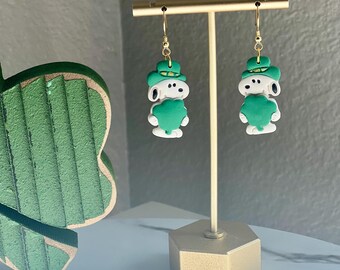 Puede incluir: Un par de pendientes de aro dorados con un perro de dibujos animados blanco que lleva un sombrero verde y sostiene un trébol verde. Los pendientes se muestran en un soporte dorado.