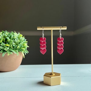 May include: A pair of dangle earrings with three stacked red heart charms. The earrings are on a gold stand.
