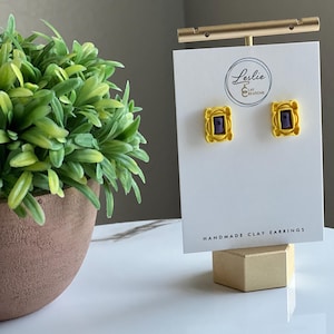 May include: A pair of yellow clay earrings shaped like the iconic purple frame from the TV show "Friends". The earrings are handmade and are displayed on a white card with the text "Leslie Ear Creations" and "Handmade Clay Earrings".