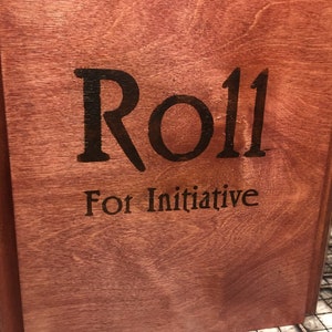 May include: A wooden box with the words "Roll for Initiative" engraved on the front. The box is stained a dark reddish brown.