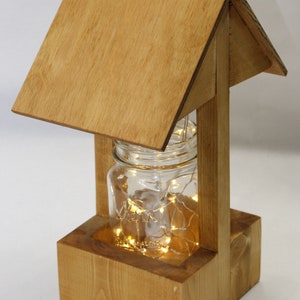 May include: A wooden wishing well-shaped decorative piece. It features a clear glass jar filled with string lights, set within a wooden frame. The frame has a pitched roof and a square base, all made of light-colored wood.