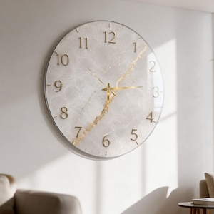 May include: A round wall clock featuring a light gray marble-effect face with a prominent diagonal gold vein running across the center. The clock has raised gold-colored numerals from one to twelve and thin, matching gold-colored hour and minute hands.