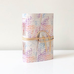 May include: A white journal with a colourful floral pattern and a tan leather strap. The journal is closed and tied with the strap.