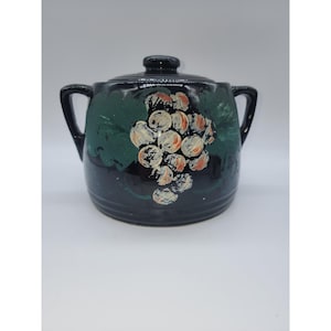 Puede incluir: Un azucarero de cerámica negra con tapa y dos asas. El cuenco está decorado con un diseño de hojas verdes y un racimo de uvas en tonos blancos, naranjas y negros. El azucarero es un artículo de menaje.