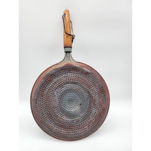 May include: A round, copper-colored metal strainer with a perforated surface and a dark, weathered patina. It features a long, cylindrical wooden handle attached with a metal ferrule and a small, thin metal loop for hanging at the end of the handle.