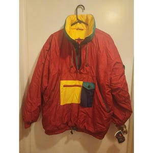 May include: A red anorak jacket with a yellow, green, and navy blue color block design. The jacket has a high collar, a front pocket, and a drawstring at the waist. The jacket is hanging on a black hanger.
