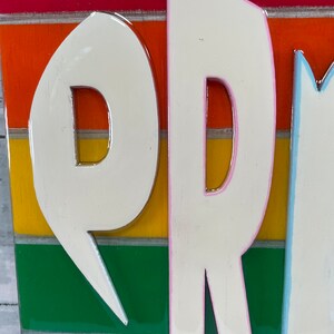 May include: Close-up of a wooden sign with the word "PR" in white raised letters with pink outlines. The letters are on a rainbow background with red, orange, yellow, green, blue, and light blue stripes.