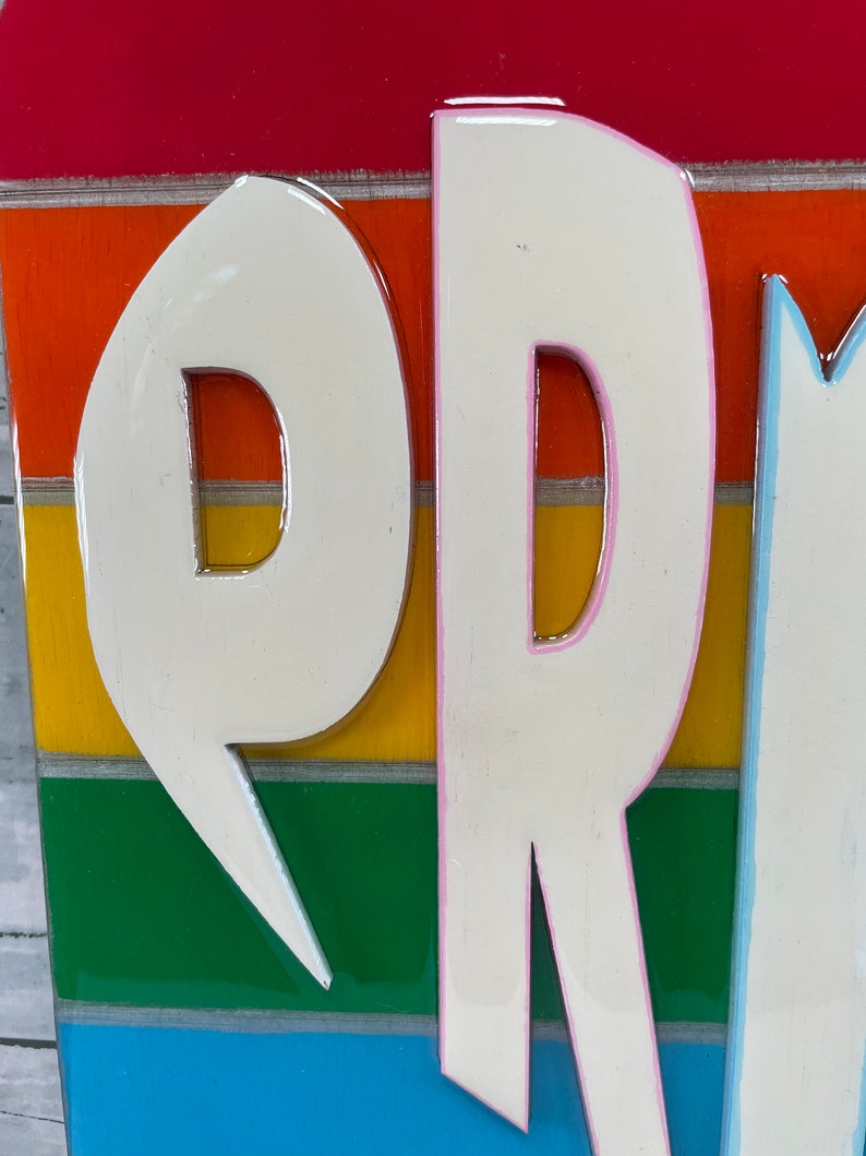 May include: Close-up of a wooden sign with the word "PR" in white raised letters with pink outlines. The letters are on a rainbow background with red, orange, yellow, green, blue, and light blue stripes.