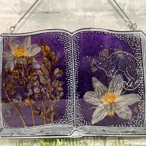May include: A purple resin book with a white outline and a pressed flower design. The book has a silver chain attached to the top and a cat silhouette on the right page.