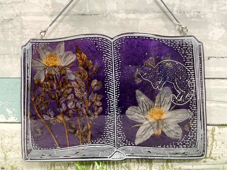 May include: A purple resin book with a white outline and a pressed flower design. The book has a silver chain attached to the top and a cat silhouette on the right page.