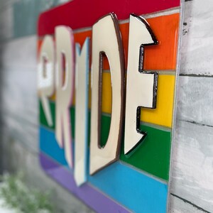 May include: A wooden sign with the word "PRIDE" in white letters with a brown outline. The letters are raised and have a 3D effect. The background is a rainbow of colors, with each stripe representing a different color of the rainbow.