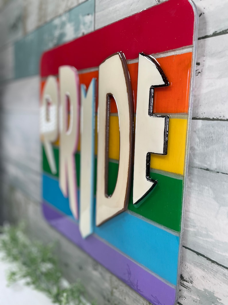 May include: A wooden sign with the word "PRIDE" in white letters with a brown outline. The letters are raised and have a 3D effect. The background is a rainbow of colors, with each stripe representing a different color of the rainbow.