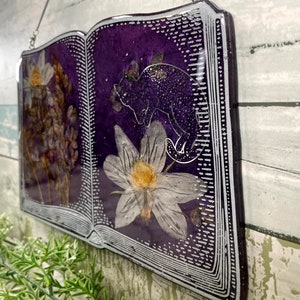 May include: A purple book-shaped resin ornament with a silver border. The book is open and features pressed flowers and a silhouette of a cat.