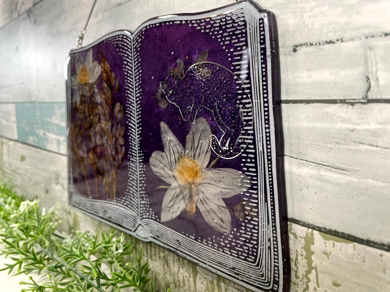 May include: A purple book-shaped resin ornament with a silver border. The book is open and features pressed flowers and a silhouette of a cat.