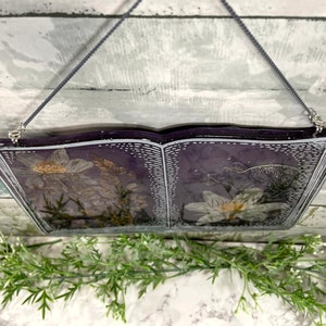 May include: A book-shaped resin wall hanging with dried flowers pressed inside. The book is open and the pages are a dark purple color. The flowers are white and have a delicate, vintage look.