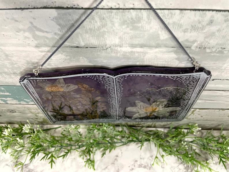 May include: A book-shaped resin wall hanging with dried flowers pressed inside. The book is open and the pages are a dark purple color. The flowers are white and have a delicate, vintage look.