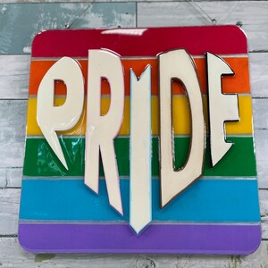 May include: A wooden sign with the word "PRIDE" in white raised letters. The sign is painted with the colors of the rainbow flag.