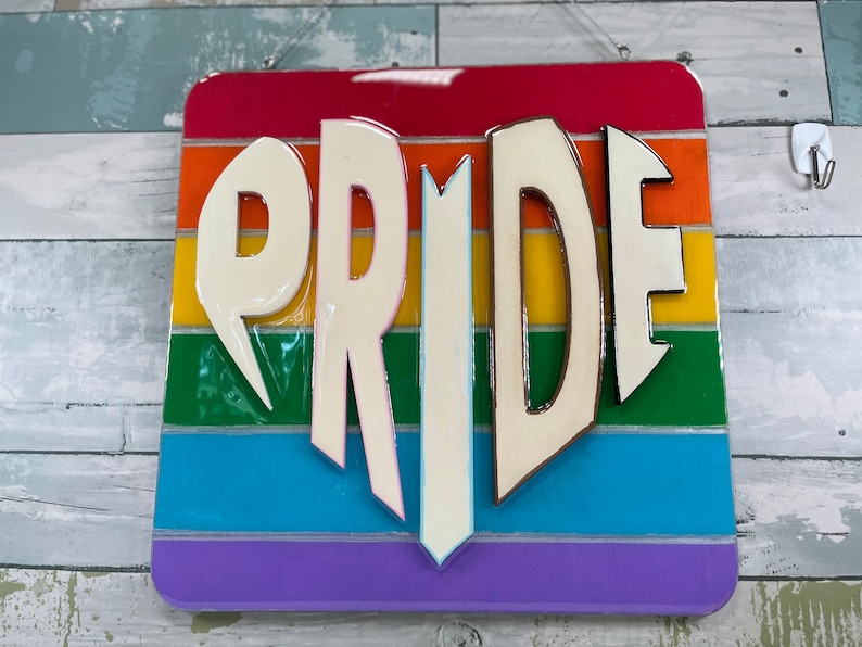 May include: A wooden sign with the word "PRIDE" in white raised letters. The sign is painted with the colors of the rainbow flag.