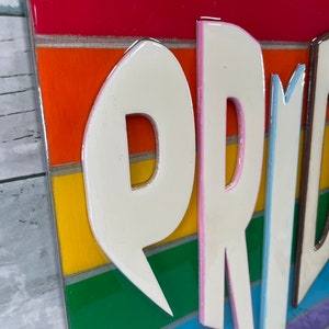 May include: A rainbow-colored wooden sign with the word "PRIDE" in white raised letters with pink and blue outlines. The letters are layered on top of the rainbow stripes.