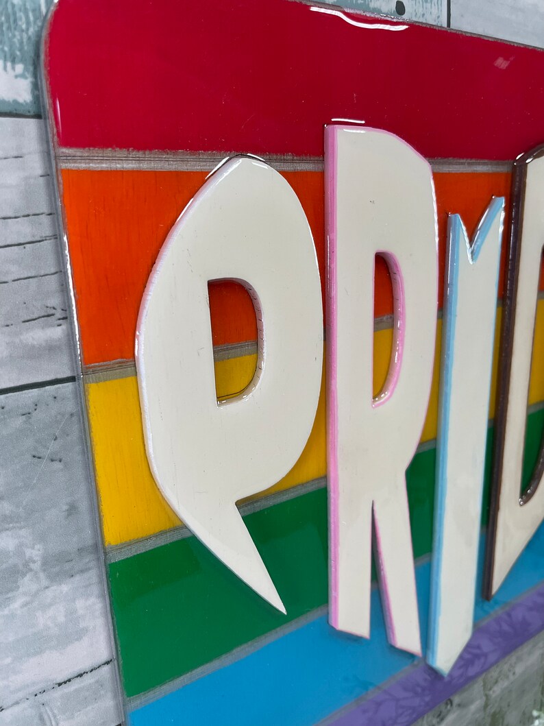 May include: A rainbow-colored wooden sign with the word "PRIDE" in white raised letters with pink and blue outlines. The letters are layered on top of the rainbow stripes.