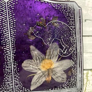 May include: A purple resin book with a white outline and a pressed flower and a silhouette of a cat inside. The book is hanging from a silver chain.
