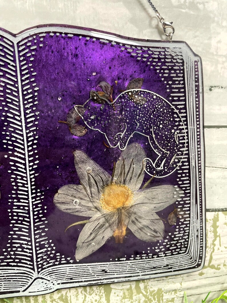 May include: A purple resin book with a white outline and a pressed flower and a silhouette of a cat inside. The book is hanging from a silver chain.