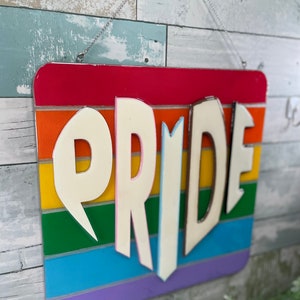May include: A rainbow-colored wooden sign with the word "PRIDE" in white letters. The sign is hanging from a silver chain.